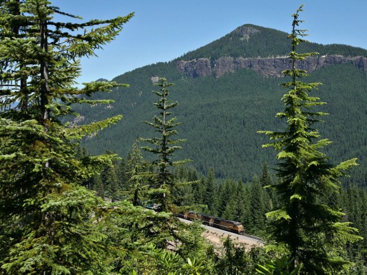 Southbound work train carrying welded rail ascending the Cascades near Cruzatte, Oregon. The rocky southern flank of Mount David Douglas appears in the background. The mountain (as well as the Douglas Fir tree) is named for a Scottish botanist who explored the Pacific Northwest in 1824.