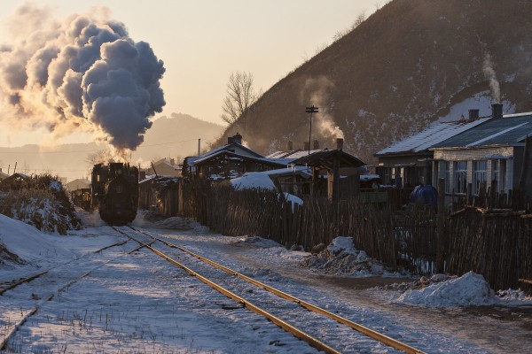 In the long shadows and warm light of a cold November afternoon, an empty coal train steams through the village of Tuoyaozi, Heilongjiang, China, on the Huanan Forestry Railway.