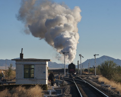 On a fall morning in Gulumanhan, Inner Mongolia, China, a westbound freight train departs behind a pair of 2-10-2 QJ steam locomotives. The switch tender at the west end of the siding waits to inspect the train as it passes.