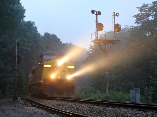 CSX eastbound coal train cuts through a foggy summer morning at Amblersburg, West Virginia, passing the color position light signals installed by predecessor Baltimore & Ohio. The eighty-car train, with two more locomotives pushing on the rear, is starting the climb up Cranberry Grade on the aptly-named Mountain Subdivision.