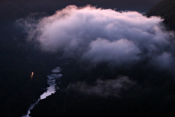 Clouds of fog follow the New River downstream as a CSX empty hopper train rolls west through the predawn darkness near Nuttallburg, West Virginia. The view is from a long cliff at the top of the New River Gorge known to rock climbers as the "Endless Wall."