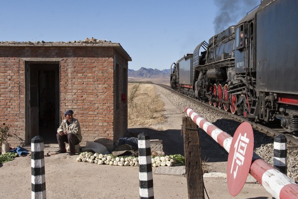 A crossing gatekeeper along the Jitong Railway near Dariqiga, Inner Mongolia, China, watches from his shanty as a westbound freight train passes behind two QJ steam locomotives in October 2005.