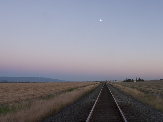 Seen from a rural road crossing between Tangent and Shedd, Oregon, Union Pacific's main line cuts an arrow-straight path south up the Willamette Valley as a half moon hangs in the evening sky of July 12, 2008. The foothills of the Cascades rise in the southeast.