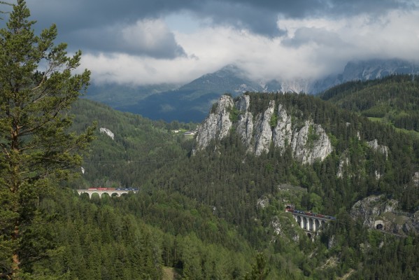 Austria's "20-Schilling view." The rocky face of the Polleroswand dominates this scene at Breitenstein, below which sits the most remarkable stretch of the Semmering Railway. At lower right is the Krauselklause Tunnel and Viaduct, with the upper level of the Kalte Rinne Viaduct visible at left, separated by the Polleroswand Tunnel. A northbound intermodal freight train stretches across both bridges and tunnels. Austria's 20 Schilling banknote from 1967 featured this scene. Today a hiking trail and a lookout tower provide easy access to this famous vantage.