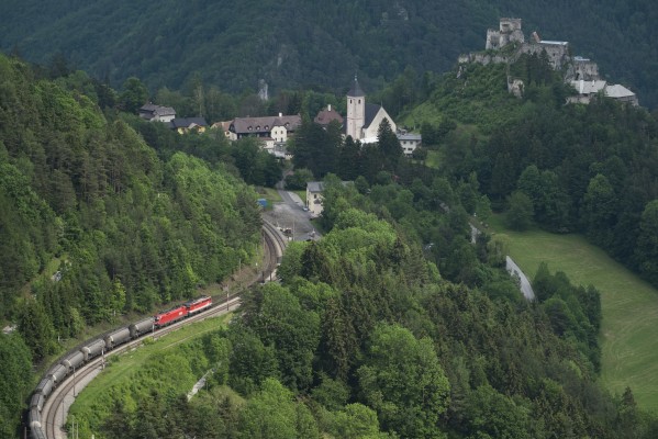 Northbound freight train winding downgrade at Klamm, passing the Catholic church and ruins of Castle Klamm, which dates from the 13th century. The cemetery in the churchyard includes an obelisk monument for the workers who died building the Semmering Railway, estimated at 1,000 men.