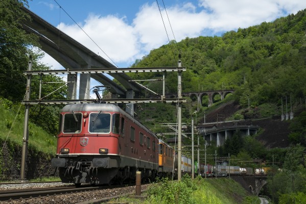 Two Swiss electrics lead an intermodal train south at Biaschina. Two additional levels of track are visible behind the train, connected by the Piano Tondo and Travi spiral tunnels. The A2 motorway, completed in 1980, soars over the scene at upper left.
