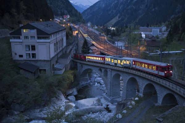 At Göschenen, the narrow-gauge Schöllenen Railway meets the Swiss Federal main line at the northern portal of the Gotthard Tunnel. At dusk, a narrow gauge train of the Matterhorn Gotthard Railway is crossing the River Reuss as it begins the rack-assisted climb to Andermatt.