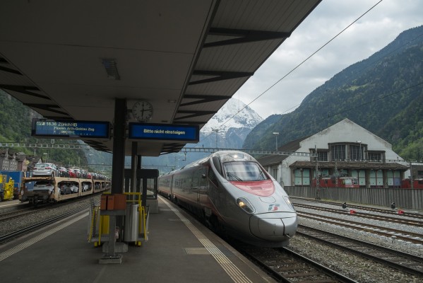 EuroCity train north from Milan to Zurich passes through Erstfeld station while a train of new automobiles waits for clearance south. At right is the enginehouse that serviced helper locomotives and today maintains a fleet of historic equipment.