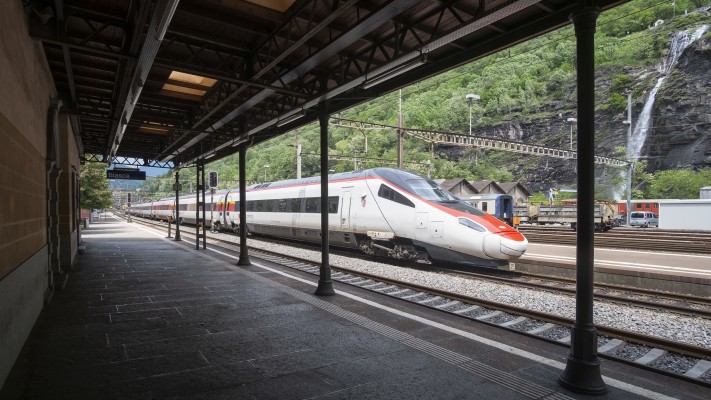 An ICN tilting train breezes through Biasca station with the Santa Petronilla waterfall in the background.