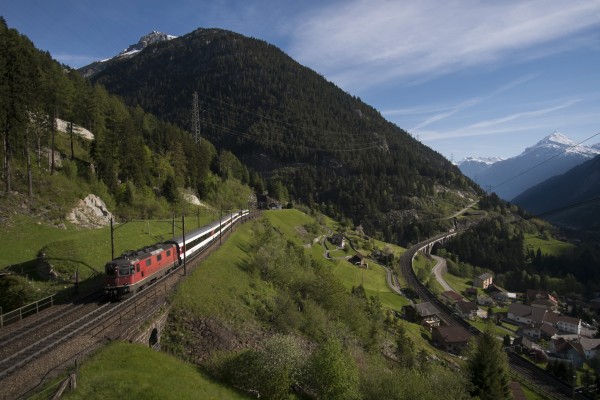 An InterRegio passenger train climbs the top level of track at Wassen, Switzerland, in brilliant morning sunshine with snow-capped Chli Windgällen towering in the distance at 9,797 feet.
