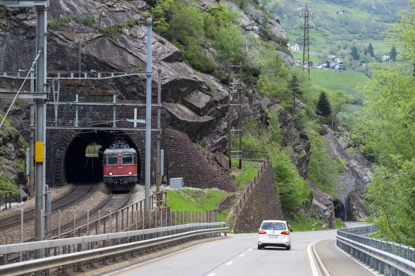 An InterRegio passenger train north emerges from the Dazio Tunnel just below Rodi-Fiesso. The road descends into the Piottino Gorge at a much sharper angle, which the railway mitigates via the Freggio spiral tunnel.
