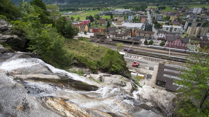 The Santa Petronilla waterfall spills down toward the Biasca station as an ICN passenger train passes.