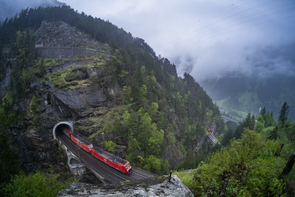 Two red Deutsche Bahn electric locomotives haul a freight train over the Upper Meienreuss Bridge above Wassen on a rainy spring morning. Only a fisheye lens can take in the full scene of this tight but spectacular vantage.