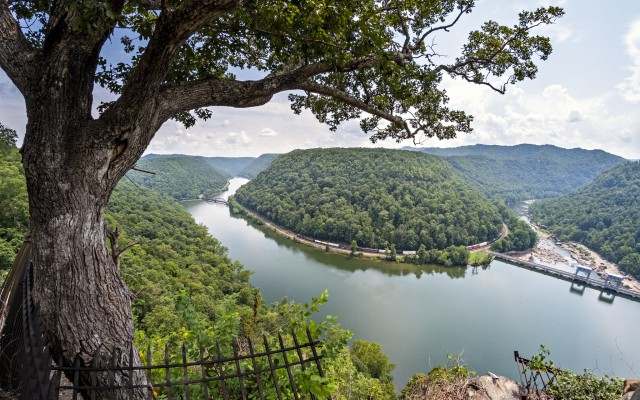 An oak tree at West Virginia’s Hawks Nest overlook frames the gorge of the New River as CSX merchandise train H750 slogs eastward on a humid August afternoon. The single AC-traction locomotive on the headend is assisted by a second unit in the middle of the train. Together, they’re making maybe fifteen miles per hour on their twisting, upstream run on Main Track Two—part of this train’s daily trek from Russell, Kentucky, to Rocky Mount, North Carolina. Just ahead, Track One diverges to cross the river on a pair of Parker through trusses. Eight miles beyond, Track Two crosses the river on a single Parker truss to rejoin Track One at Sewell. Before this train finishes passing through the scene, a westward empty hopper train will ease across the bridge—a flurry of activity for a line that currently sees about a dozen trains a day.
For all the beauty it offers, Hawks Nest has a tragic history. The location takes its name from osprey that used to nest in the cliffs here. Blasting during railroad construction from 1869 to 1873 disturbed the birds and they never returned, although you can still see vultures catching thermals at eye level. Most of the New’s flow exits the gorge via a tunnel next to the dam at lower right, which runs three miles under Gauley Mountain to provide hydroelectric power for a metals plant at Alloy. Constructed in the late 1920s and early 1930s, the tunnel runs through nearly pure deposits of silica. Project managers demanded grueling hours and provided almost no safety equipment, and hundreds of the workers who dug the tunnel later died from silicosis.
Hawks Nest is a touchstone for me. As a frequent stop on family outings during my childhood, it is the place where the notion of the railroad in the landscape first enthralled me. In my early days as a photographer I visited frequently, making a few memorable images and more than a few memories. One of my favorite visits was a summer morning in 2007 with my grandfather, when there were no trains to disturb our conversation. As I learned more of the history of Hawks Nest, it has become a place to try to reconcile conflicting feelings over technology and its price. Even as life has taken me far from Hawks Nest, it remains a powerful memory, a thought that never fails to elicit strong emotions.
This visit was a bonus, a detour and a brief stop on the way to meet part of my wife’s family, and my first time here in more than a decade. We stayed for less than half an hour, and you could easily wait hours here without seeing any trains. Somehow we saw two. (Although it's hard for me to think of an eastward merchandise train as anything other than Q302...) Even in the high backlight of a summer afternoon, all of the magic was still there. For me, I suspect it always will be.