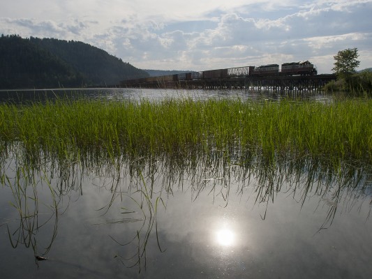 Still waters at the edge of Benewah Lake reflect the late afternoon July sun as the St. Maries River Railroad's inbound Plummer Turn returns from the Union Pacific interchange at Plummer, Idaho, with empty boxcars and flatcars for the Potlatch Mill in St. Maries.